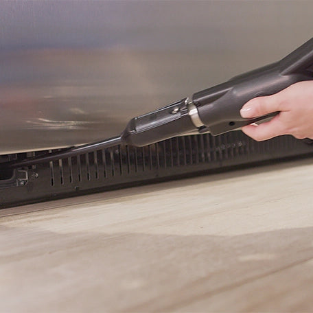 Person using coil cleaner attachment on  the bottom of a refrigerator. 