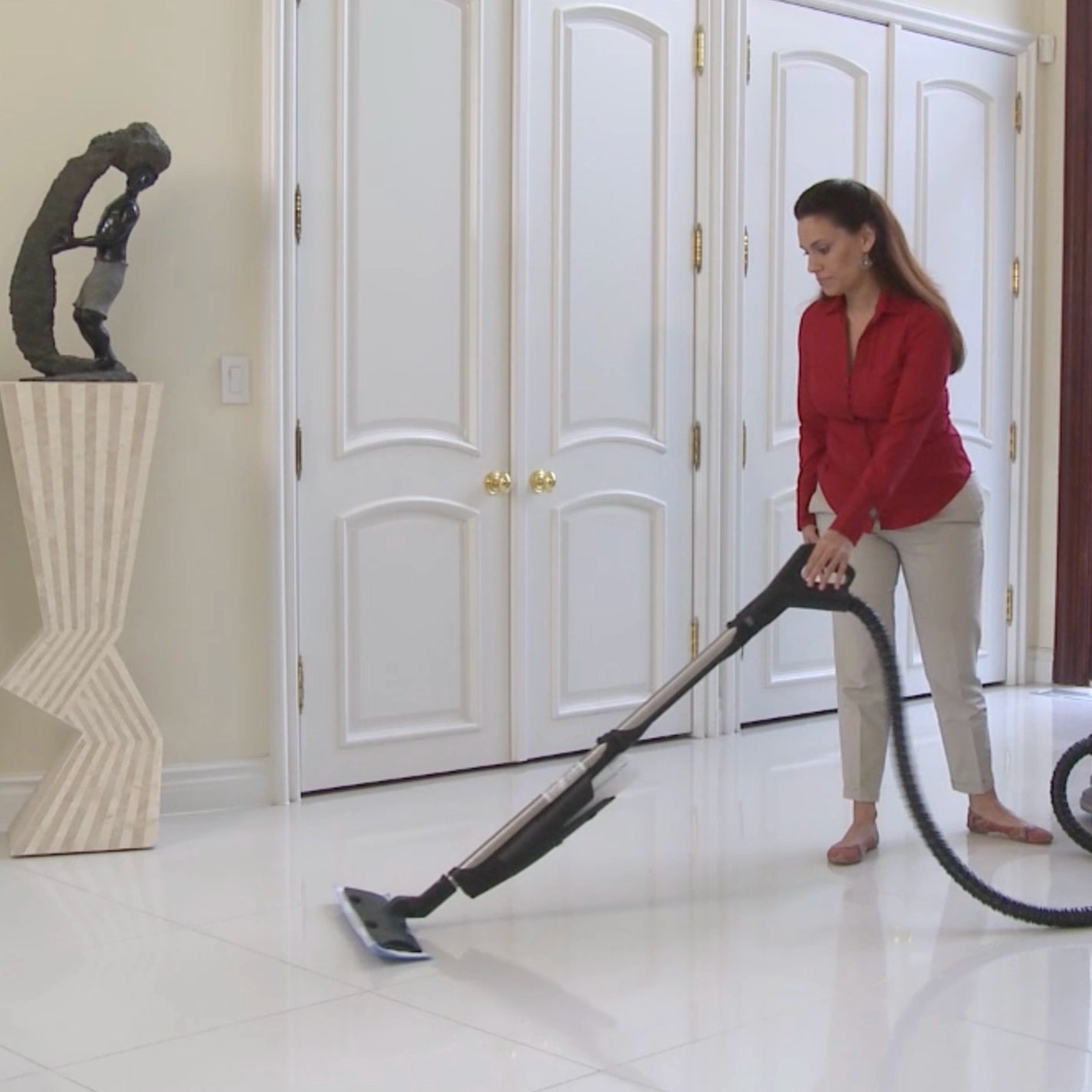 Woman cleaning a white tiled floor with a Rainbow vacuum cleaner with JetPad attachment in a room with white doors and a sculpture.